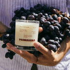 Hand holding the Earl of East Uva scented candle in front of a bunch of red grapes